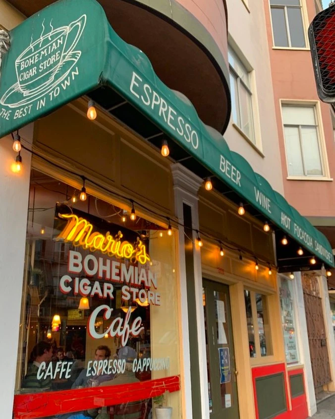 Exterior de Mario's Bohemian Cigar Store Cafe en San Francisco dónde se ve el toldo verde con una taza de café dibujada y un cartel de neón amarillo con el nombre del local detrás del vidrio