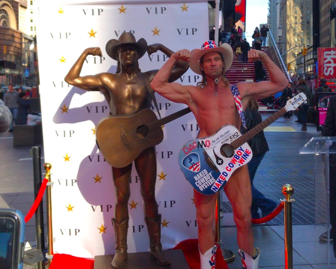 Naked Cowboy de Times Square posando junto a su estatua frente a las escaleras rojas de Manhattan