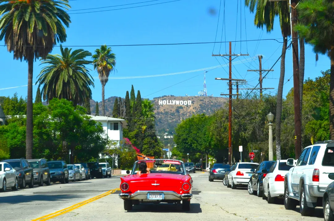 Auto rojo andando sobre una calle de Los Ángeles con el cartel de hollywood al forndo