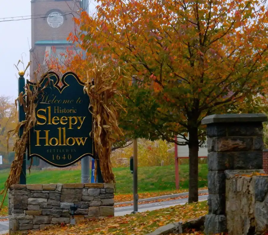 _pueblo_ny_sleepy... Cartel de entrada a Sleepy Hollow con un árbol de hojas naranjas en otroño y la torre de la iglesia detrás